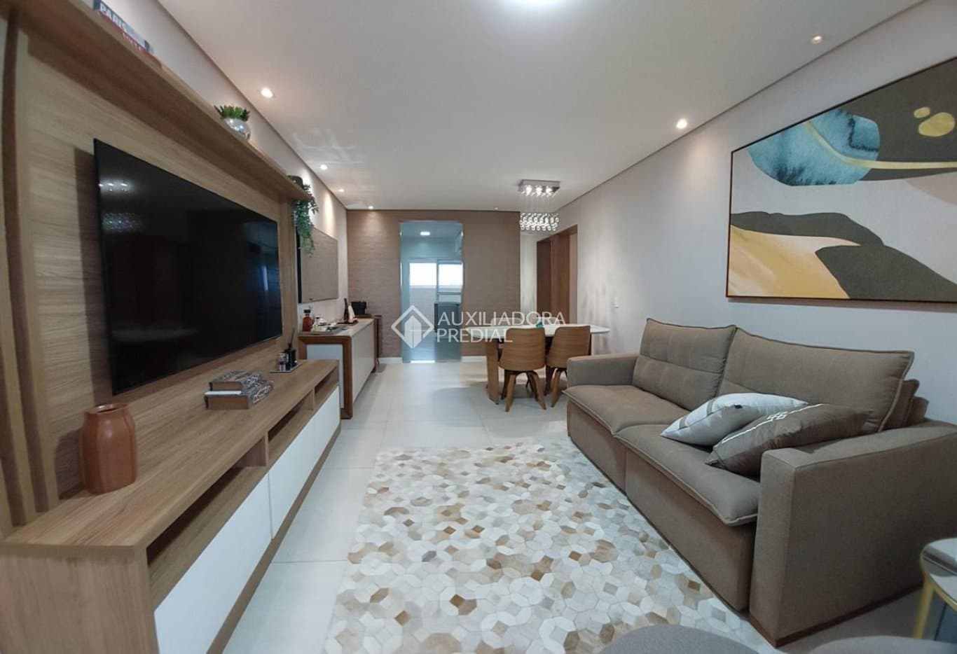 Sala de estar com painel para TV, sofá marrom, tapete estampado e mesa de jantar ao fundo.